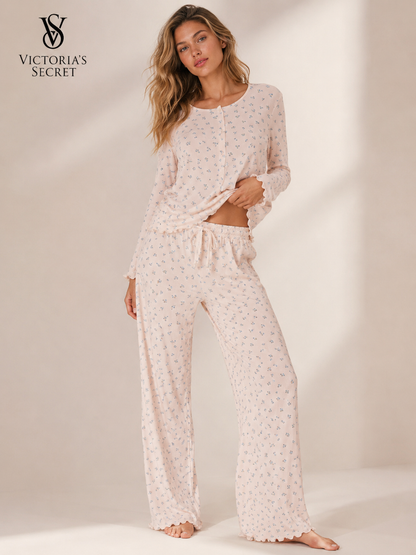 A woman modeling a light pink long-sleeve pajama set with a small floral print, featuring a long-sleeve top and matching wide-leg pants, shown against a warm beige studio background with the Victoria's Secret logo in the top left corner.