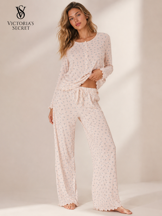 A woman modeling a light pink long-sleeve pajama set with a small floral print, featuring a long-sleeve top and matching wide-leg pants, shown against a warm beige studio background with the Victoria's Secret logo in the top left corner.